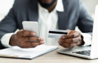 man at desk holding smartphone and credit card (Depositphotos)
